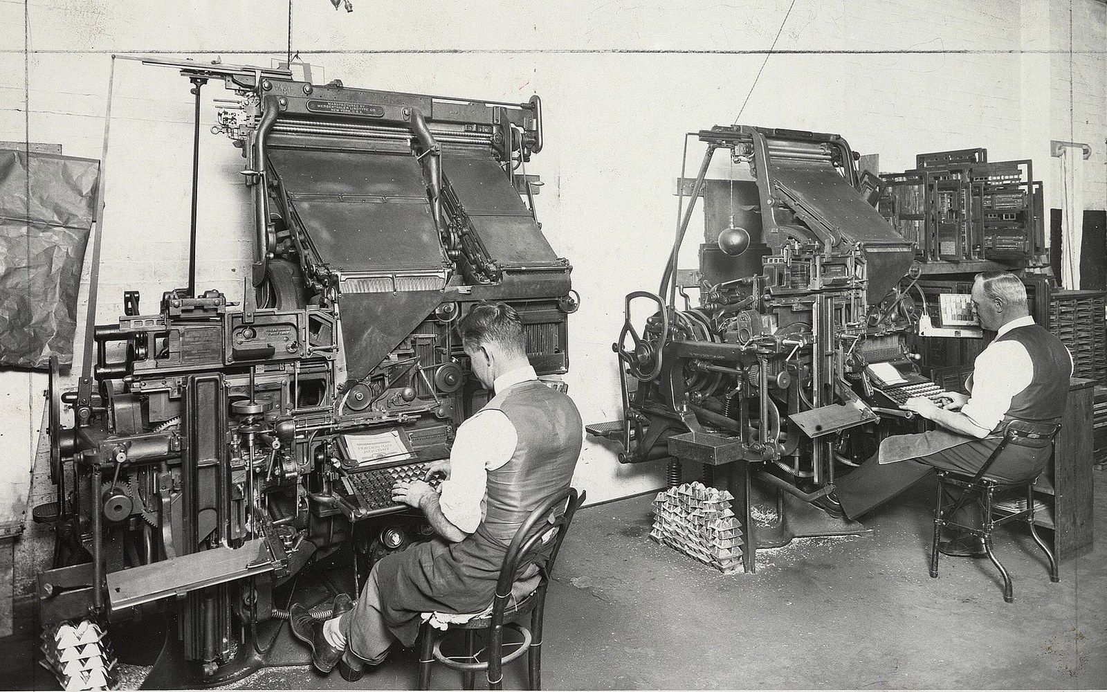 Two typesetters operate large Linotype machines at their keyboards in a print shop
